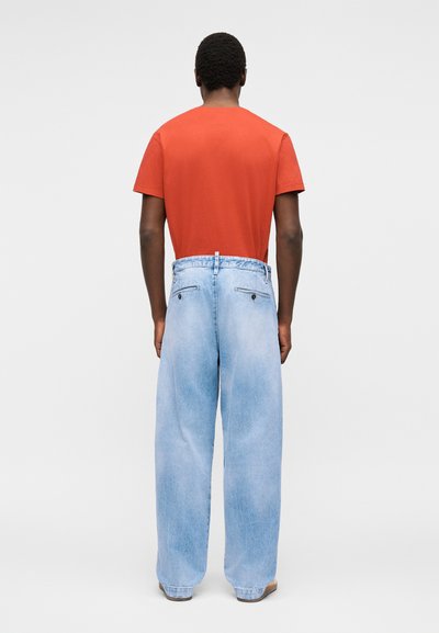 Άνδρας που φοράει φαρδιά ανοιχτομπλέ τζιν και απλό πορτοκαλί κοντομάνικο μπλουζάκι, στέκεται με την πλάτη σε απλό φόντο.