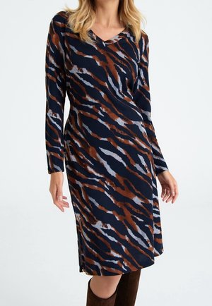 Vrouw draagt een navyblauwe midi-jurk met lange mouwen met bruine en grijze abstracte tijgerstrepen, gecombineerd met bruine kniehoge laarzen.