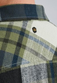 Chemise en flanelle à carreaux avec des motifs verts, bleus et blancs. Présente une étiquette noire avec du texte doré sur le col. Tissu texturé.