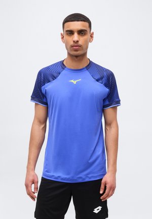 Man die een blauw sportshirt met zwarte patroonmouwen en een zwarte korte broek draagt, staand tegen een effen achtergrond.