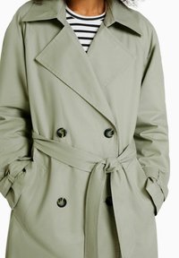 Světle olivově zelený trenčkot s dvouřadým zapínáním, velkým límcem, stahovacími pásky v pase a černými knoflíky. Nosit na pruhovaném topu.