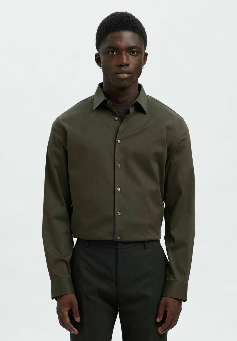 Ciemnozielona koszula z długim rękawem i kołnierzykiem zapinanym na guziki, z delikatną, paskowaną fakturą i pasującymi guzikami. Noszona z ciemnymi spodniami.