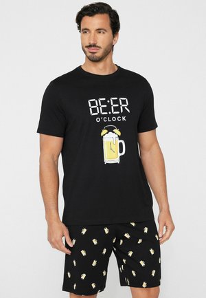 Homme portant un t-shirt noir avec le texte "BEER O'CLOCK" et un graphisme d'horloge avec une chope de bière, associé à un short noir à motifs de chopes de bière.
