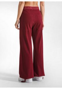 Bordeaux brede broek van een zacht materiaal, met een elastische tailleband met de tekst "GODDESS" en twee achterzakken. Gecombineerd met witte sneakers.