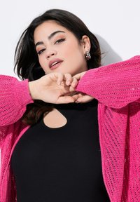 Cardigan a maglia rosa con texture a rete aperta, indossato sopra un top aderente nero con scollatura a chiave. La luce naturale mette in risalto il design.