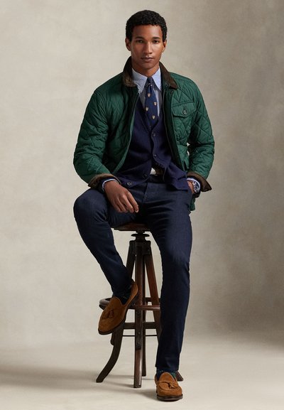 Grønn quiltet jakke med brun krage, blå genservest, stripete skjorte, mørke jeans og brune mokkasinsko, sittende på en treskammel.