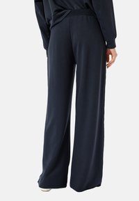 Marineblauwe wijde broek gemaakt van zachte stof, met een smocktail en een relaxte pasvorm. De broek heeft een gladde textuur en subtiele zijsplitten.
