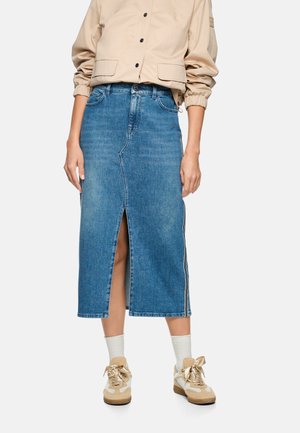 Person trägt eine beige Jacke, einen blauen Jeans-Midirock mit vorderem Schlitz, weiße Socken und beige Sneakers mit Satinbändern vor weißem Hintergrund.