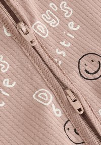 Närbild av beige ribbad tyg med dragkedja och tryckt vit text samt svarta smileys på textilen.