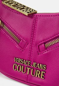 Bright pink leather bag featuring two gold zippered pockets and a gold logo plate that reads "Versace Jeans Couture." Textured finish.