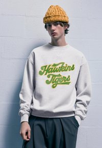 Felpa grigia chiaro con "Hawkins Tigers" in corsivo verde e giallo, abbinata a pantaloni scuri a pieghe e a un berretto di maglina testurizzato color senape.