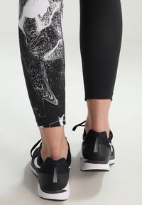 Leggings stampati in bianco e nero con un motivo a vortice, abbinati a scarpe da ginnastica nere con accenti riflettenti e suola bianca.