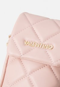 Mala de mão em pele rosa acolchoada, com um logo dourado "Valentino", costuras visíveis e uma forma triangular estruturada na parte superior.