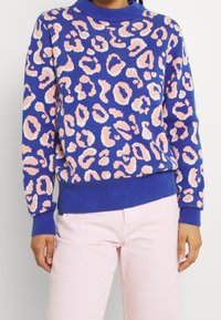 Pull imprimé léopard en bleu, avec des taches roses et blanches, poignets et ourlet côtelés, à col rond. Porté avec un pantalon rose.