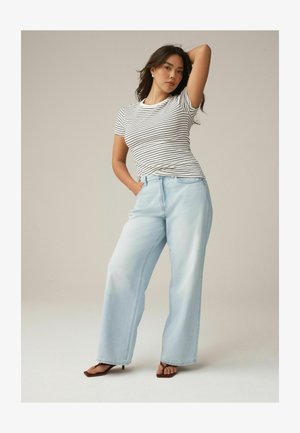 Žena s tmavými vlasmi, ktorá má oblečené bielo-čierne pruhované tričko, svetlomodré široké džínsy a hnedé sandále na opätku, pózuje s jednou zdvihnutou rukou.