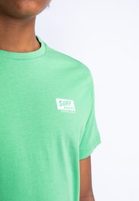 Zelené bavlněné tričko s kulatým výstřihem a krátkými rukávy, s bílou potiskem loga "SURF RIDERS PETROL IND." na hrudi.