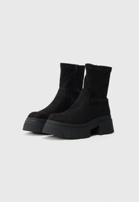 Črni semiš gležnjarji z debelim podplatom, na strani z zadrgo in gladko teksturo. Minimalistična zasnova brez vidnih znamk.