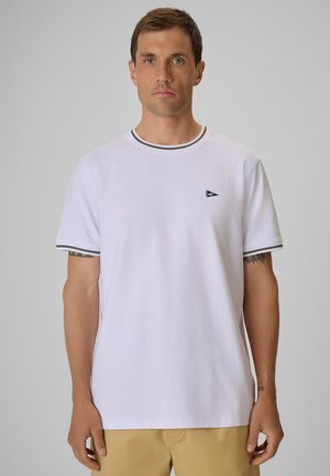 Mężczyzna w gładkim białym t-shircie z krótkim rękawem, czarnym prążkowanym kołnierzykiem i brzegami rękawów, beżowe spodnie, stojący na jasnoszarym tle.