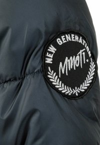 Svart nylonjacka med en slät textur, som har en rund broderad patch med texten "NEW GENERATION MMOTI" och en lagerdesign.