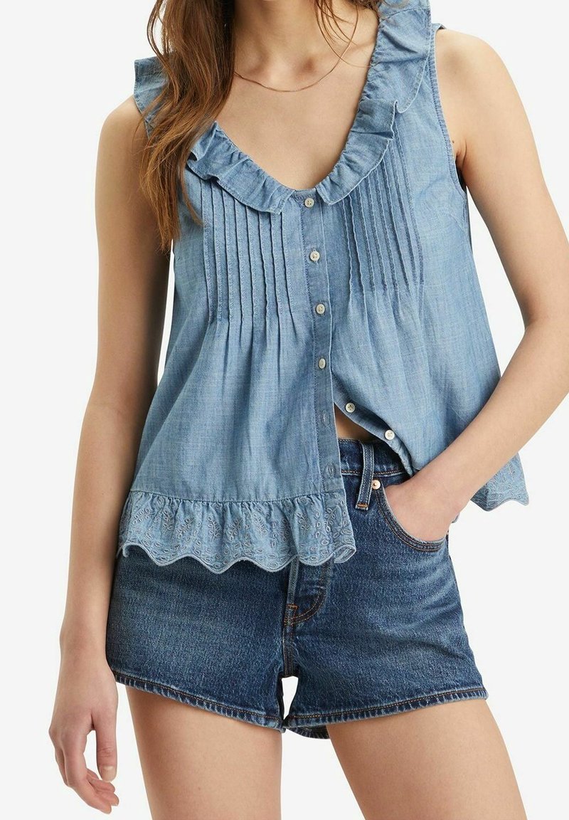 Kvinna som bär en ärmlös ljusblå knäppt topp med volanger och mörkblå jeansshorts med handen i fickan.