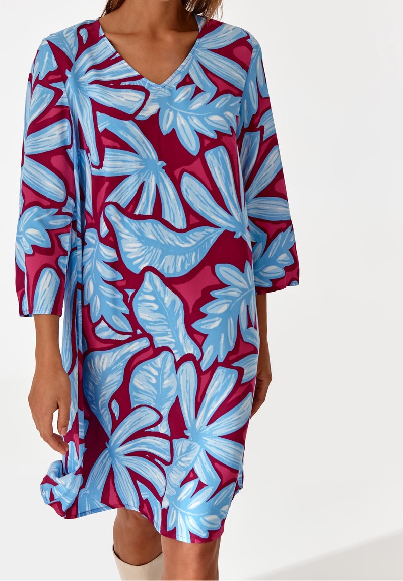 Femme portant une robe mi-longue à manches 3/4, ornée de grandes feuilles tropicales bleues sur un fond rose foncé.