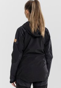 Black waterproof jacket with a hood. Features contrast fabric on sleeves, adjustable hem, and a removable brand patch on the upper arm.
