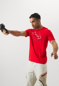 Rød T-shirt med korte ærmer og et sort Under Armour-logo, parret med lyse træningsbukser med lynlåse og detaljer.