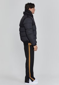 Schwarze Daunenjacke mit horizontalen Segmenten und hohem Kragen, kombiniert mit schwarzen Hosen, die einen braunen Seitenstreifen haben, und weißen Sneakers.