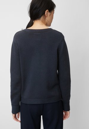 Frau mit schwarzen Haaren zu einem Pferdeschwanz gebunden, trägt einen dunkelgrauen Strickpullover und dunkle Hosen, steht mit dem Rücken zu einem schlichten hellen Hintergrund.