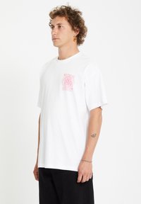 White cotton t-shirt featuring a pink spiral logo on the left chest, short sleeves, and a relaxed fit. Paired with black pants.
