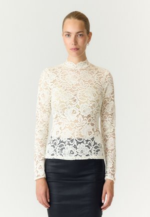 Femme aux cheveux attachés, portant un haut blanc en dentelle florale à manches longues et une jupe en cuir noire, debout devant un fond uni.