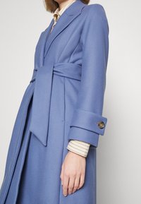 Femme portant un manteau en laine bleu clair ceinturé sur un chemisier crème à fines rayures horizontales, se tenant devant un fond uni.