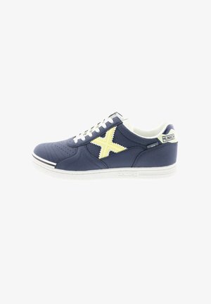 Zapatilla deportiva azul marino con detalles perforados, suela blanca, logo amarillo en forma de "X" y cordones blancos. Parte superior de tela texturizada.