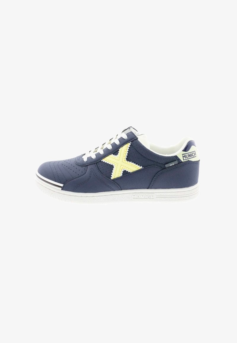 Zapatilla deportiva azul marino con detalles perforados, suela blanca, logo amarillo en forma de "X" y cordones blancos. Parte superior de tela texturizada.