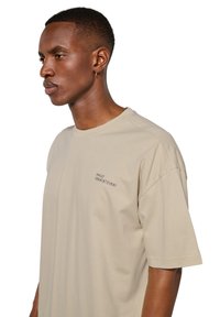 Jeune homme aux cheveux courts et petit anneau d'oreille porte un t-shirt beige oversize avec un petit texte imprimé sur la poitrine.