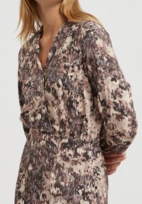 Blommönstrad klänning i dämpade nyanser av brunt, kräm och grått. Har en v-ringning och långa ärmar, tillverkad av lättviktig tyg.