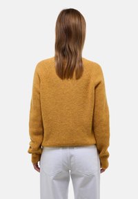 Pull en tricot jaune moutarde avec une finition texturée, doté de manches raglan et d'une coupe décontractée, associé à un pantalon blanc taille haute.