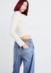 Crème kleur langemouw cropped top gecombineerd met lichtblauwe high-waisted denim jeans, met een bruine leren Lee logo patch op de tailleband.
