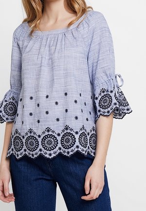Lichtblauwe blouse met zwarte bloemrijke borduurwerk langs zoom en mouwen, met 3/4 klokmouwen met strikjes, gedragen met donkerblauwe spijkerbroek.