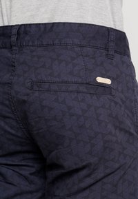Primo piano di pantaloni blu scuro con un piccolo motivo a triangolo e una tasca posteriore orizzontale, indossati con una camicia grigia chiara.
