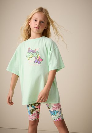 Fille blonde aux cheveux lâchés porte un t-shirt oversize vert menthe "Good Vibes" et un short coloré à motifs, debout devant un fond beige.