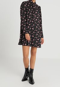 Mujer vestida con un vestido largo de manga larga negro con flores y un escote con lazo, usando botas de tobillo negras y brillantes, de pie sobre un fondo simple.