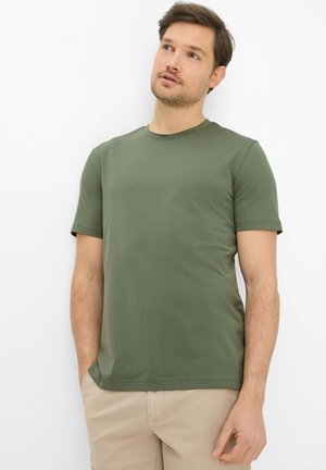 Uomo con maglietta a maniche corte verde oliva e pantaloni beige, guarda leggermente verso l'alto con le mani nelle tasche su sfondo bianco.