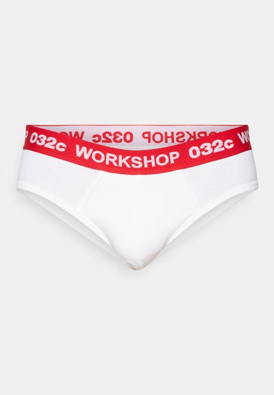 Sous-vêtements blancs avec une ceinture élastique rouge portant "WORKSHOP 032c" en lettres épaisses. Tissu lisse avec un design ajusté.