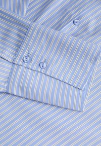 Détail d'une chemise rayée bleu clair, avec des lignes blanches verticales, un tissu lisse et deux boutons assortis sur le poignet.