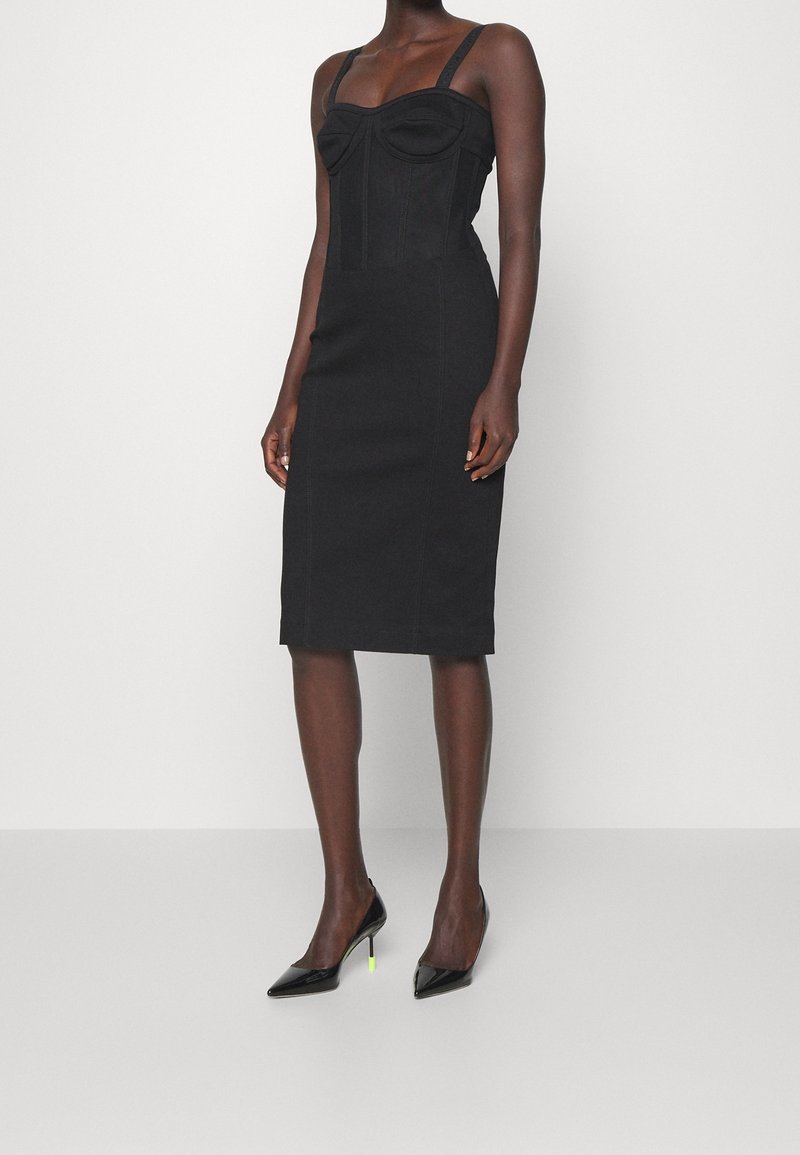 Svart figursydd klänning med en strukturerad byst, tunna band och en midjelång fåll. Har sömnadsdetaljer och är matchad med svarta högklackade skor.