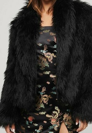 Vrouw die een zwart faux fur jasje draagt over een zwarte jurk met kleurrijke geborduurde draak- en bloemenpatronen en een dijsplit.
