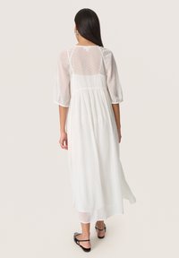 White, polka-dotted maxi dress featuring a sheer overlay, puff sleeves, and a gathered waist. Smooth fabric with a relaxed fit throughout.