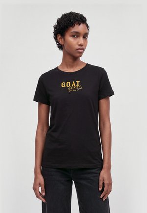 Un mannequin aux cheveux courts et bouclés porte un tee-shirt noir portant l’inscription « G.O.A.T. Le meilleur de tous les temps » et un jean foncé, sur un fond clair uni.
