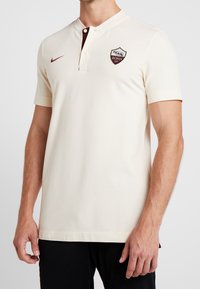 Kremowa koszulka polo z krótkim zapięciem na guziki, bordowymi akcentami i naszywką z logo Roma na lewej piersi. Miękka, gładka tkanina.
